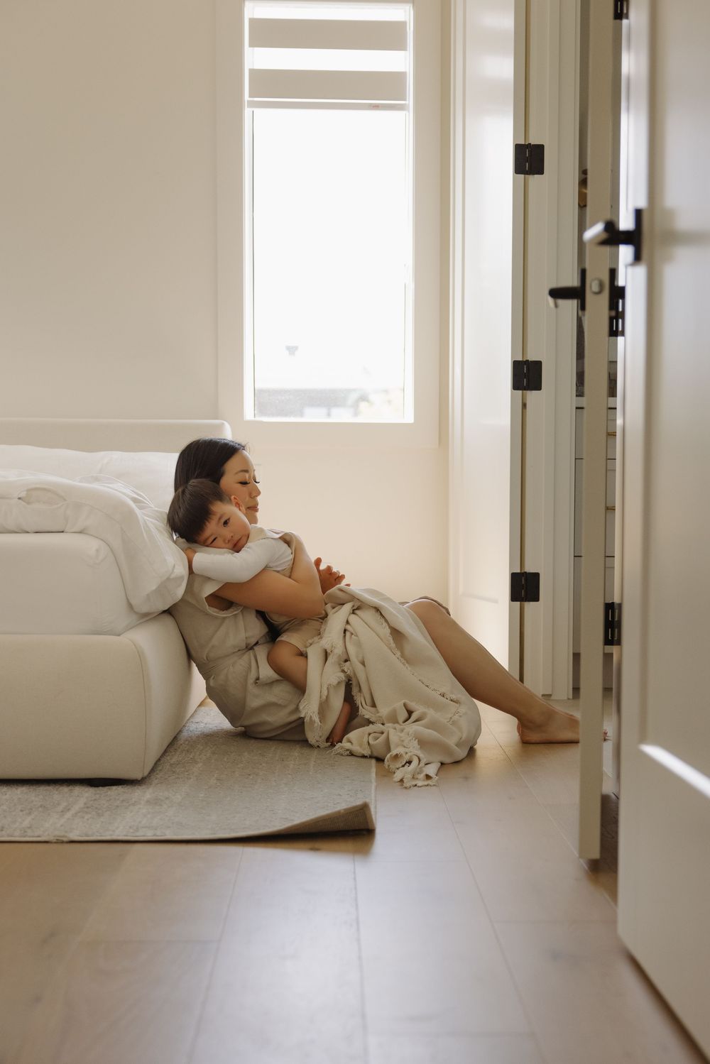 A mother sits on the floor leaning against a white sofa, tenderly hugging her young child in a bright living room.