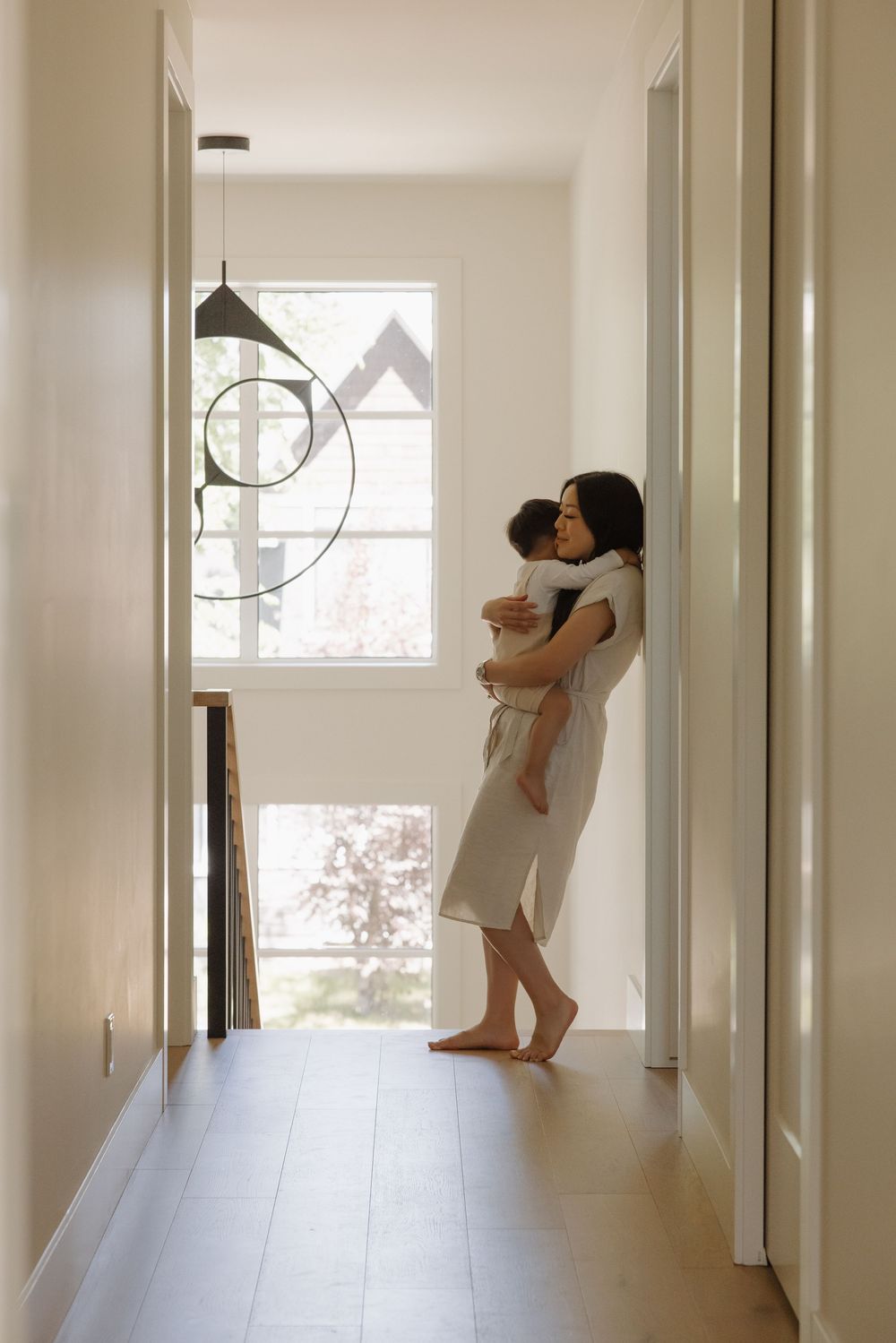 A barefoot mother tenderly holds her baby while standing in a bright modern hallway near a sunlit window.