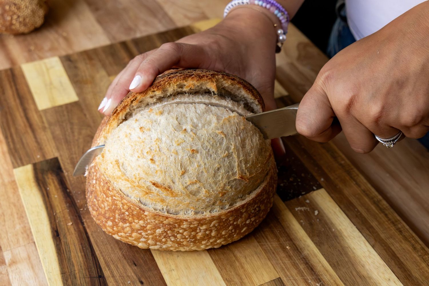 Hands slicing a rustic sourdough bread loaf on a wooden cutting board with a sharp knife.