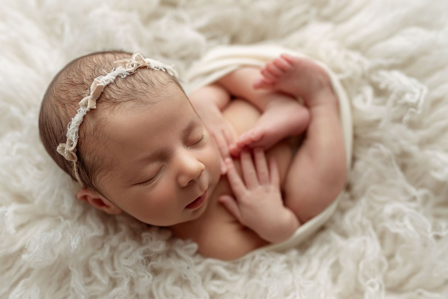Peaceful newborn baby girl sleeping on white fluffy blanket wearing a delicate lace headband.