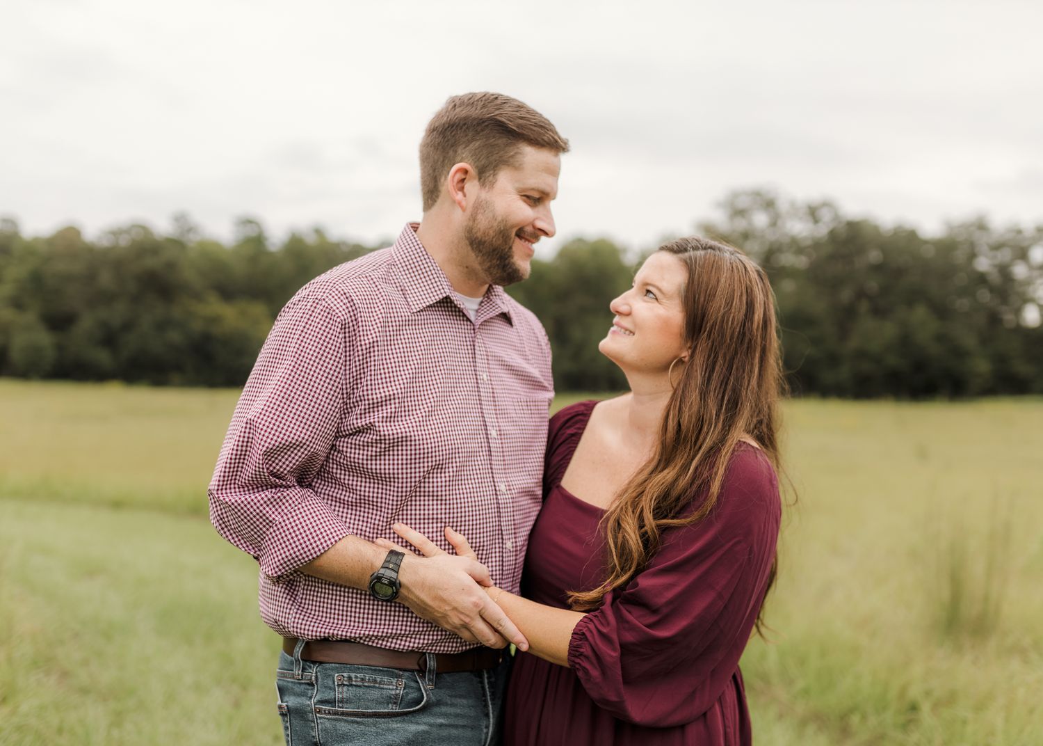 A couple holds hands walking through a lush green field surrounded by trees during an outdoor photo session.