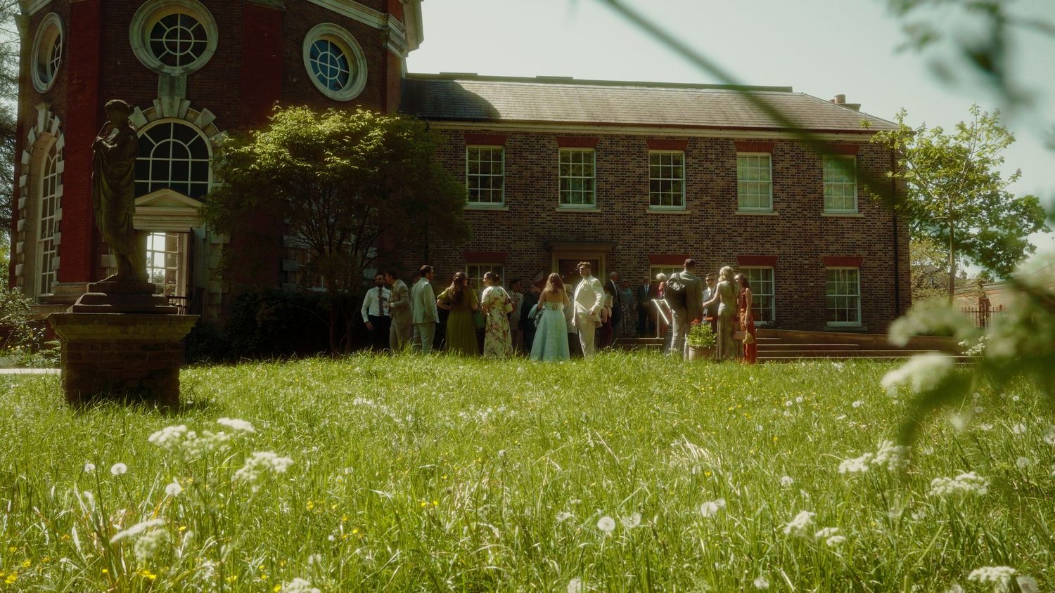 Group of people gathered outside a historic red brick building surrounded by lush green meadow with wildflowers on a sunny day.