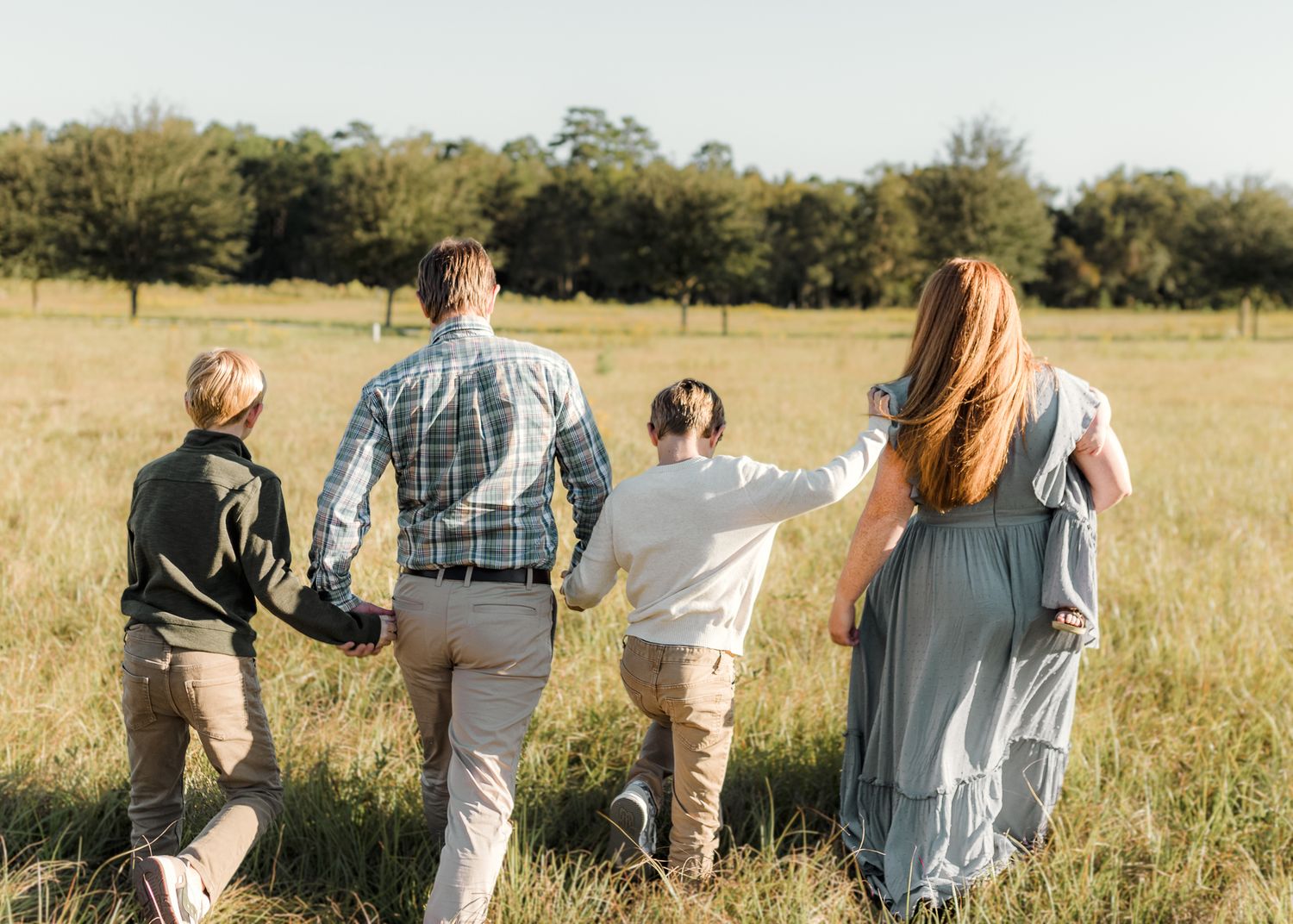 Family of five walking hand-in-hand through a sunny green meadow with trees in the background.