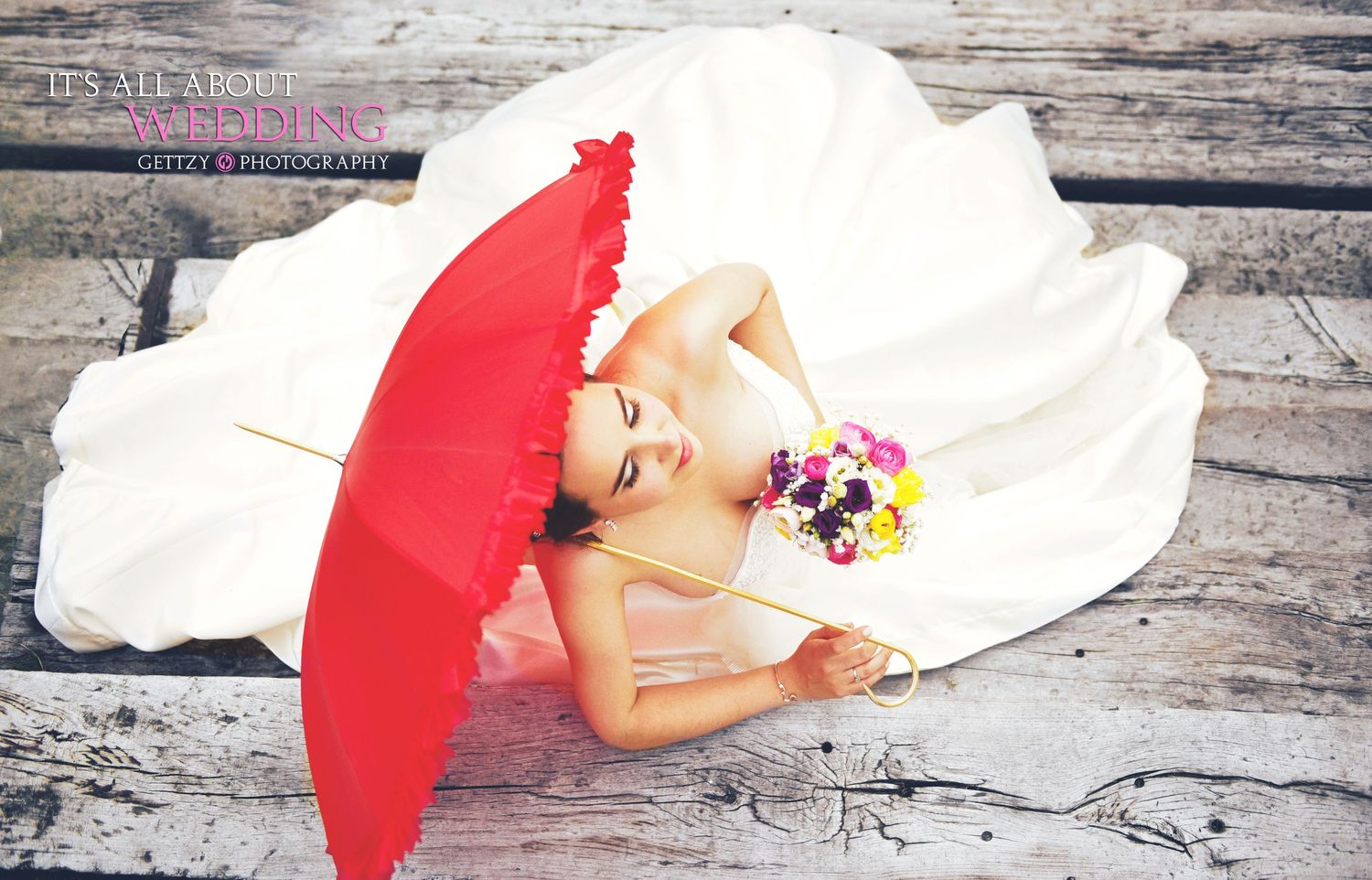 Person in a white wedding dress holding a red umbrella on wooden backdrop.