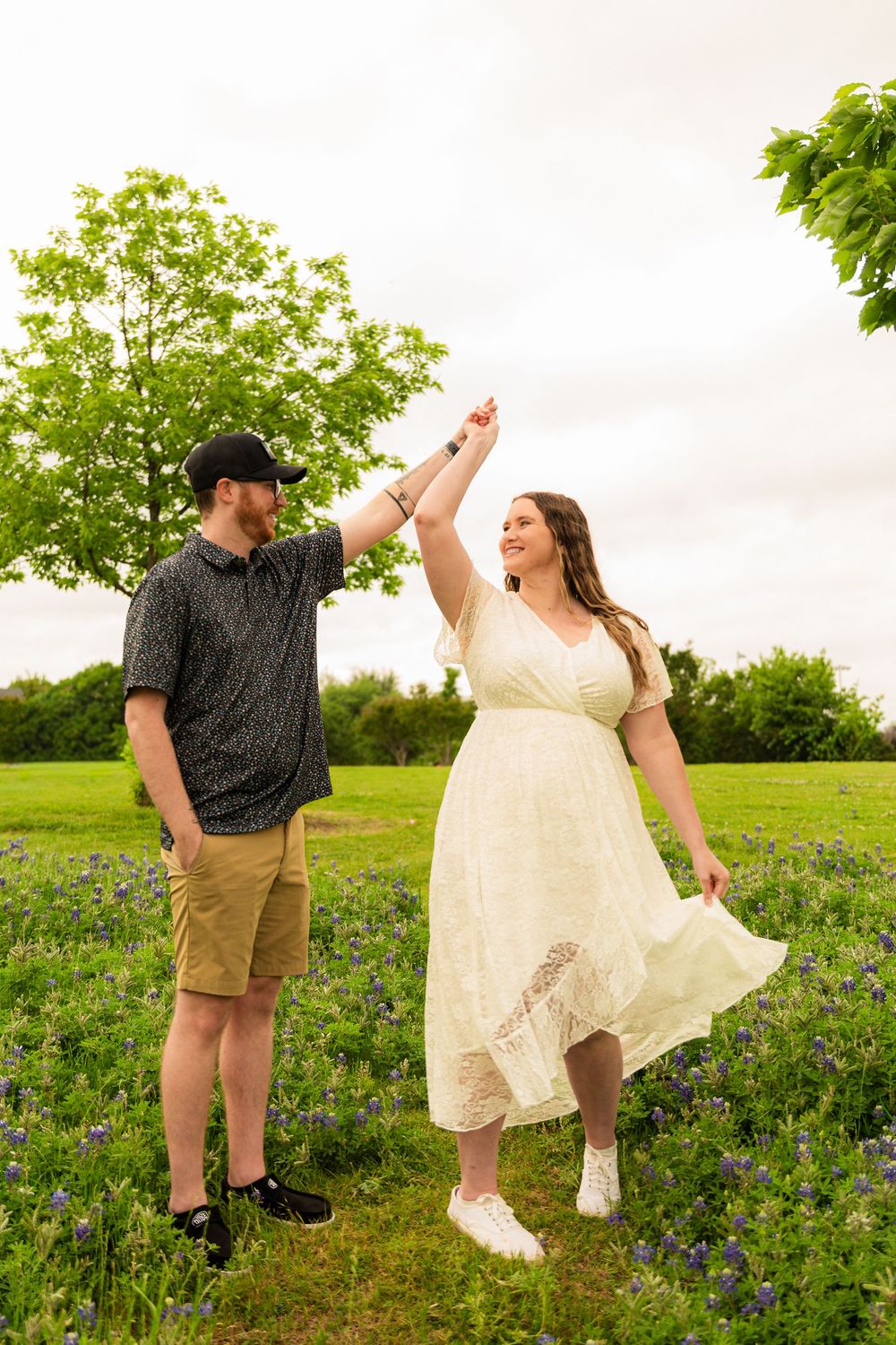 Couple dancing in a bluebonnet field, man in black cap spinning woman in white lace dress, green trees behind.