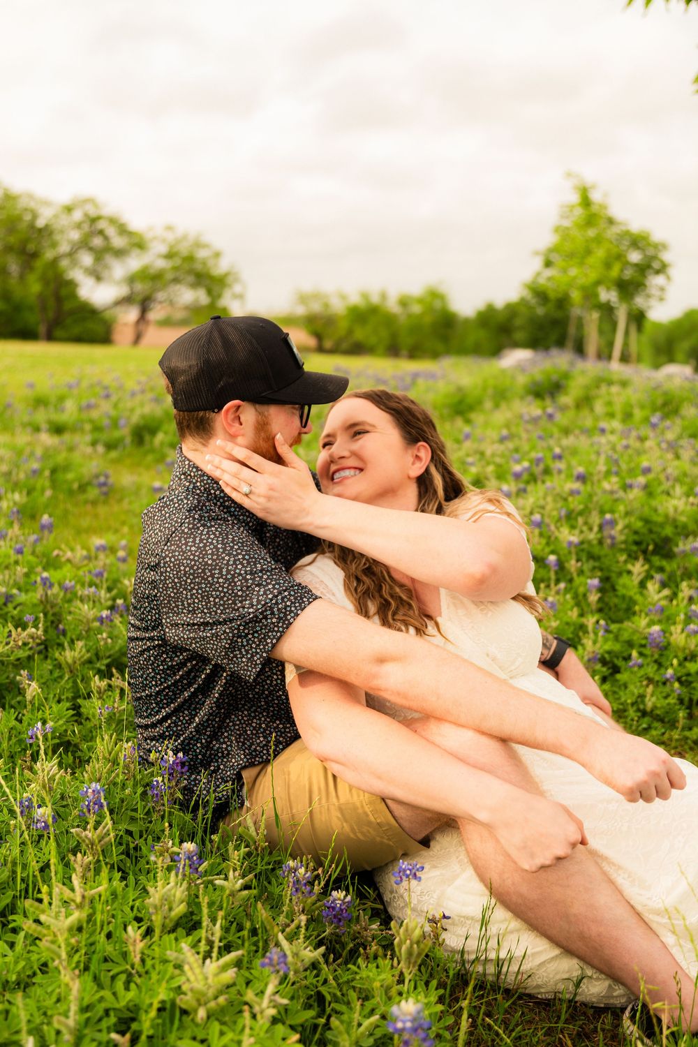 Couple sitting in a field of bluebonnets, sharing a romantic moment while the woman smiles joyfully.