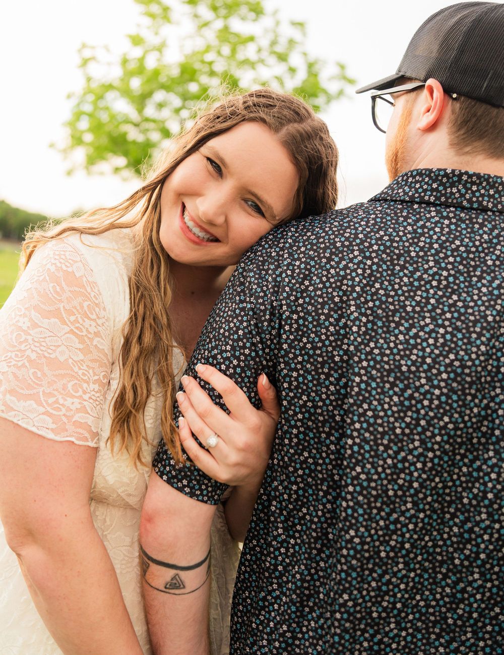 Smiling woman in lace dress hugs man in floral shirt outdoors, showing engagement ring and triangle tattoo.
