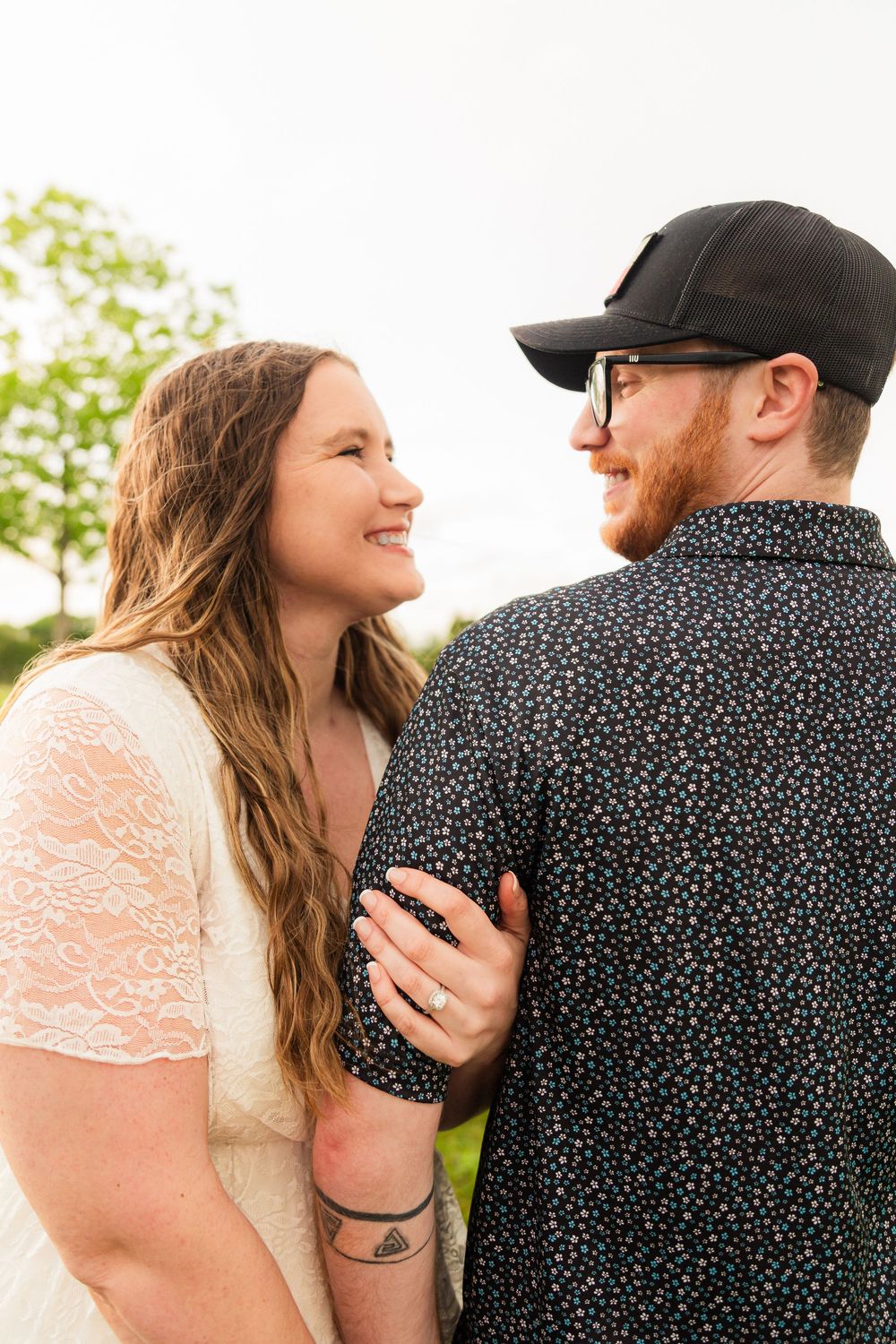 Smiling couple gazing at each other outdoors, woman in white lace dress with arm tattoo.