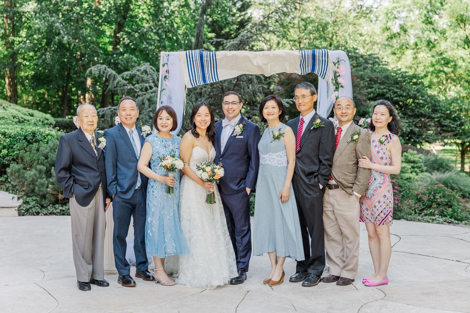 Wedding party poses outdoors under floral chuppah, bride in white gown with groom and family in elegant attire.