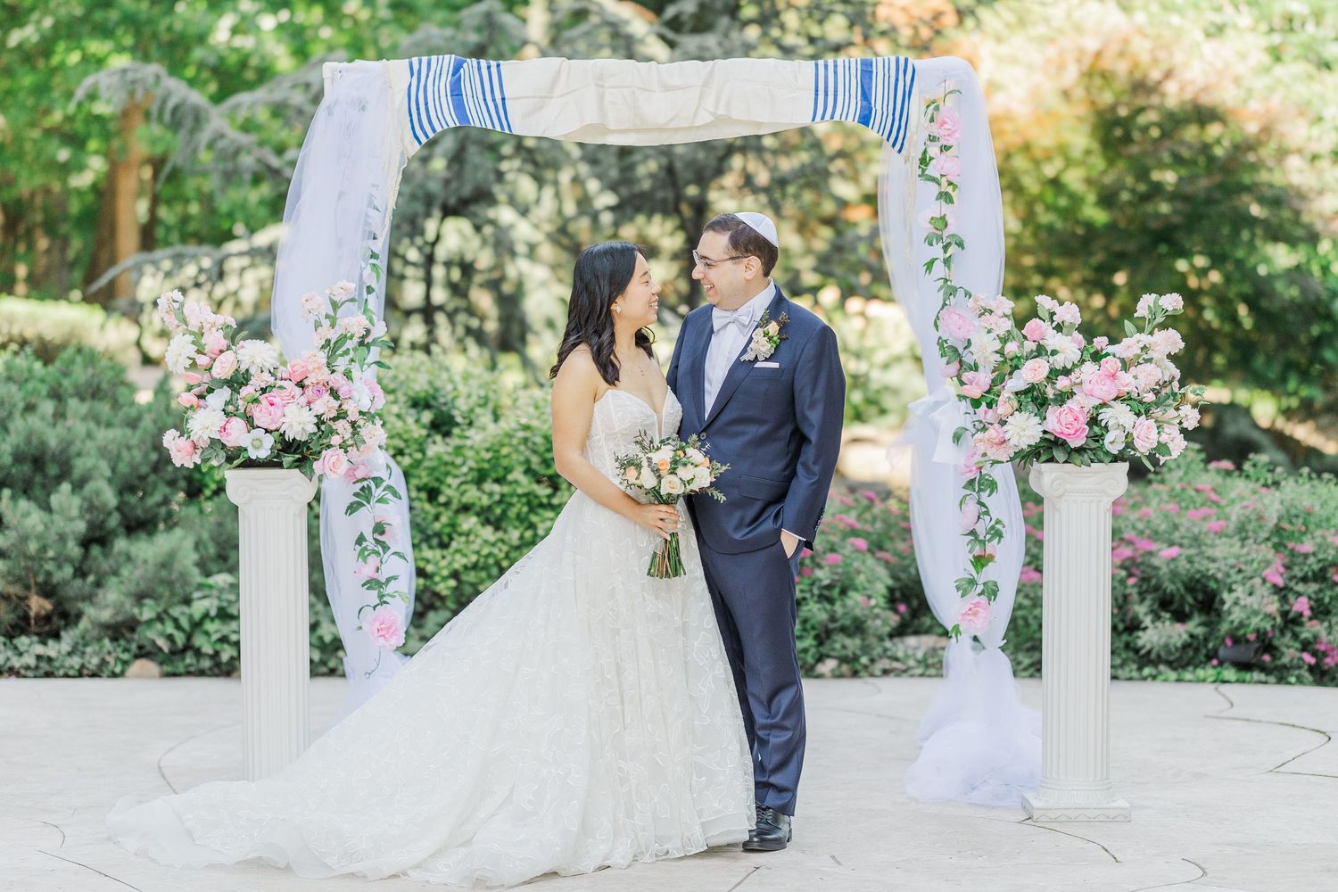 Bride and groom share a romantic moment under a floral chuppah with pink roses and greenery at an outdoor wedding ceremony.