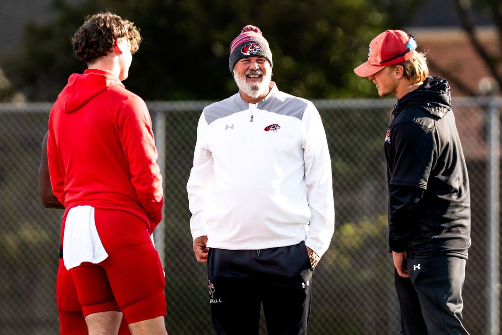 Three football coaches in red and white uniforms having a discussion on an outdoor practice field.