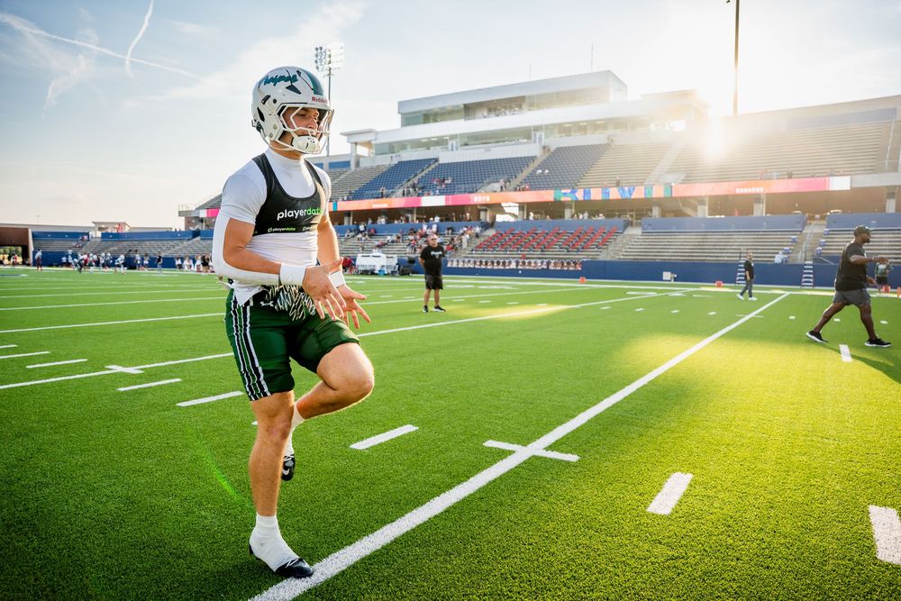 Football player in green helmet and uniform jogging on turf field during golden hour warmups.