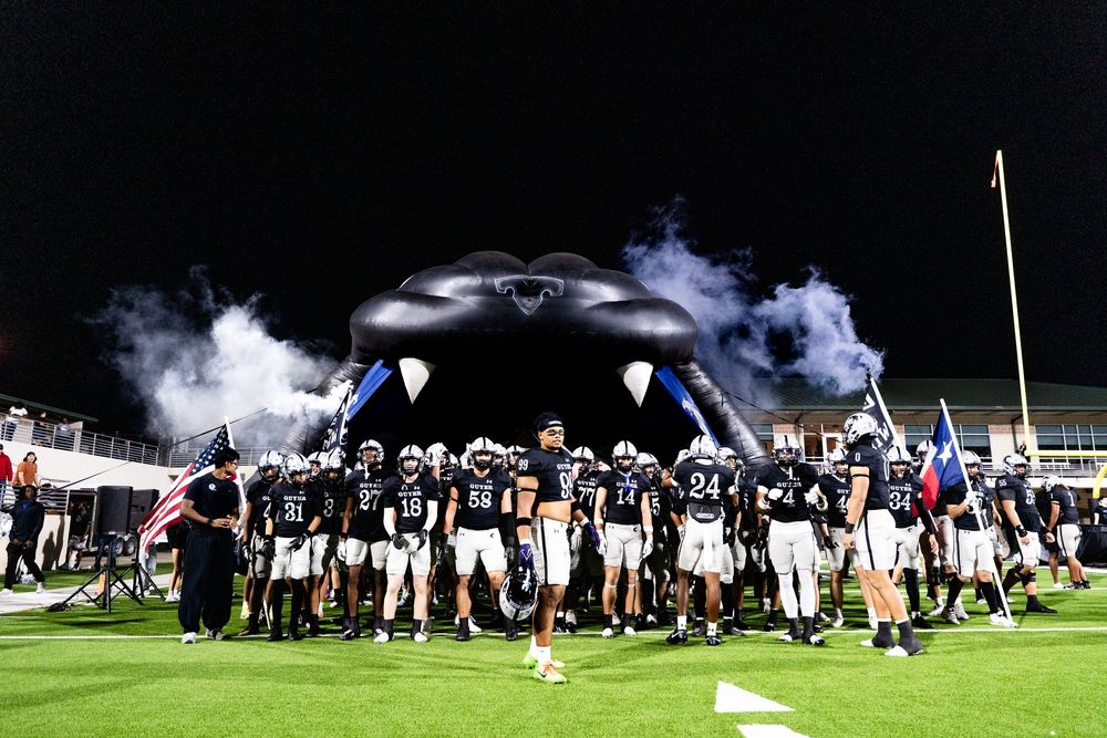 Football team entering stadium through smoke-filled inflatable tunnel under bright lights at night game.