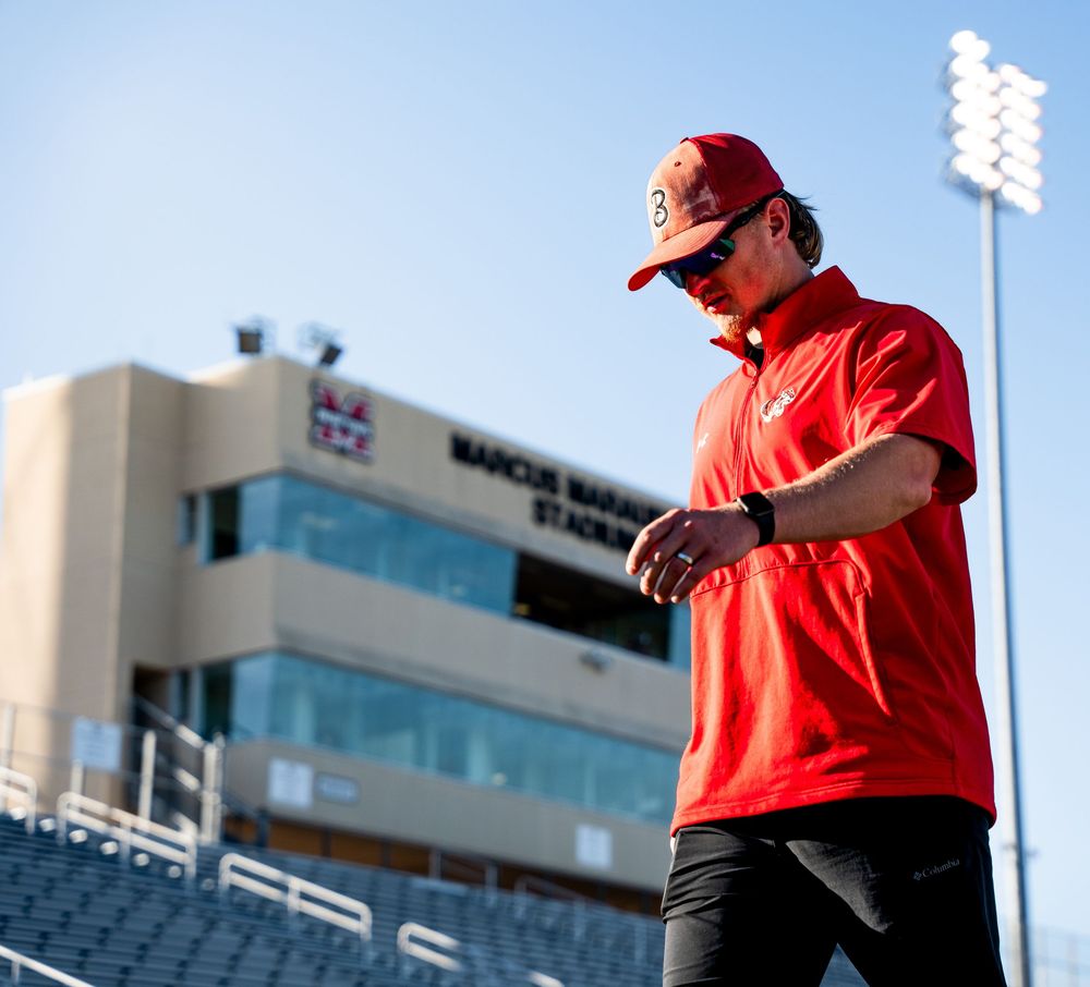 Coach in red polo shirt walking outside Marcus High School Stadium on a sunny day.