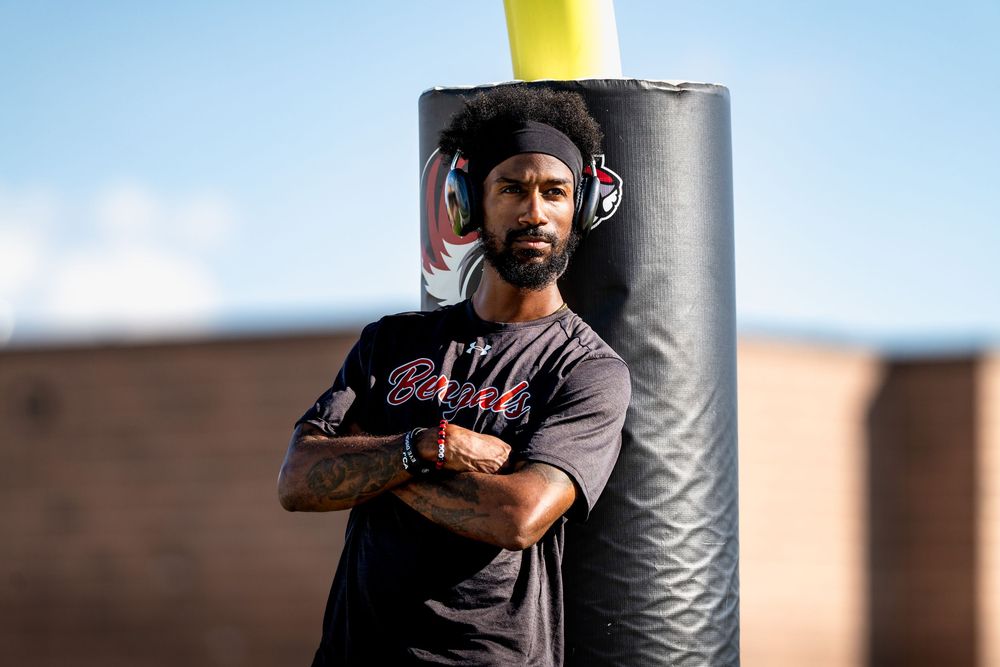 Athletic man with headphones leans against a punching bag outdoors in a black shirt.