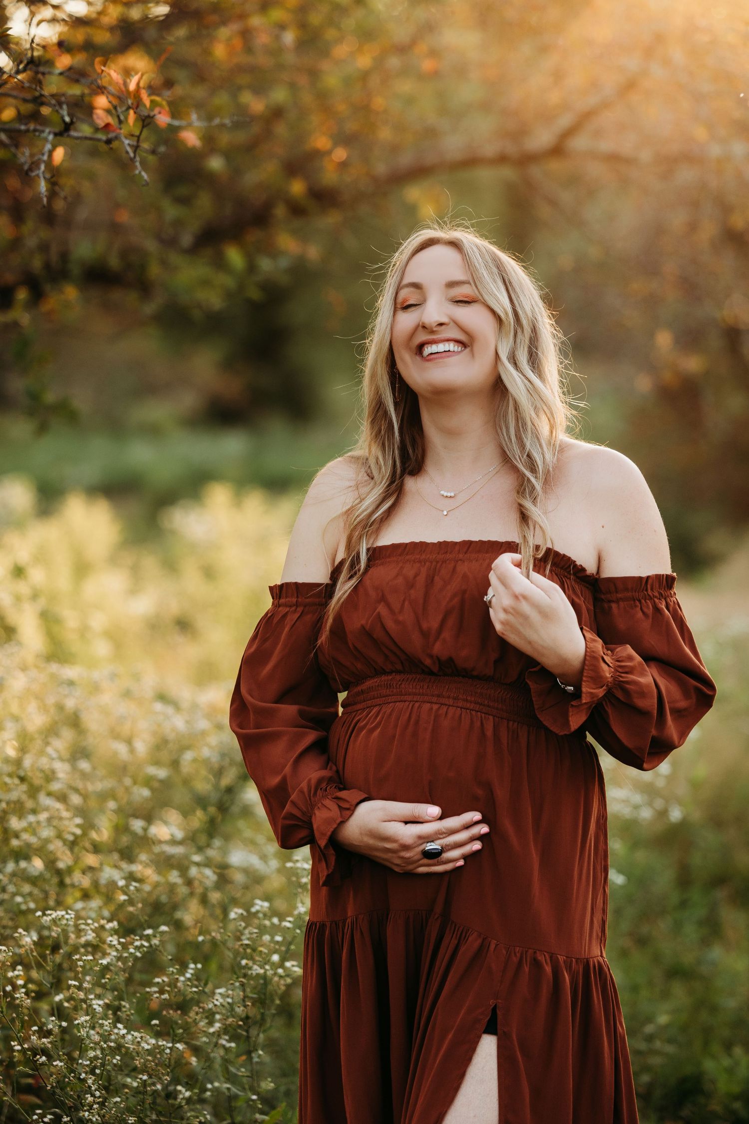 Smiling pregnant woman in rust off-shoulder dress holding baby bump outdoors in golden sunlight among wildflowers.