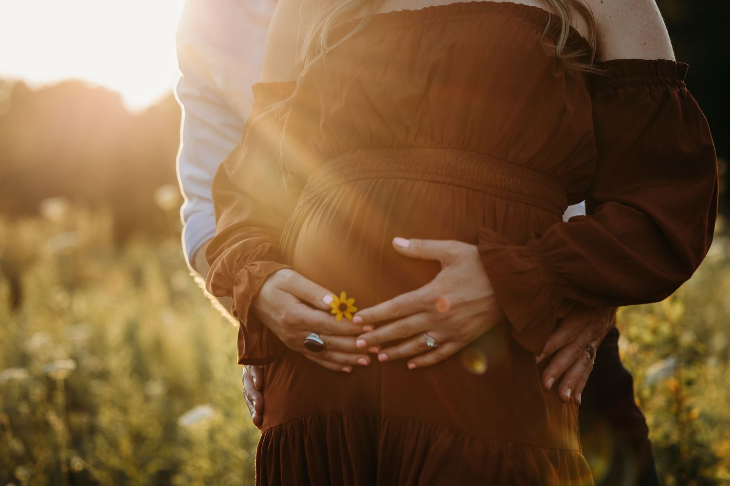 Couple embracing outdoors at golden hour, hands cradling pregnant belly in a sunlit flower field.