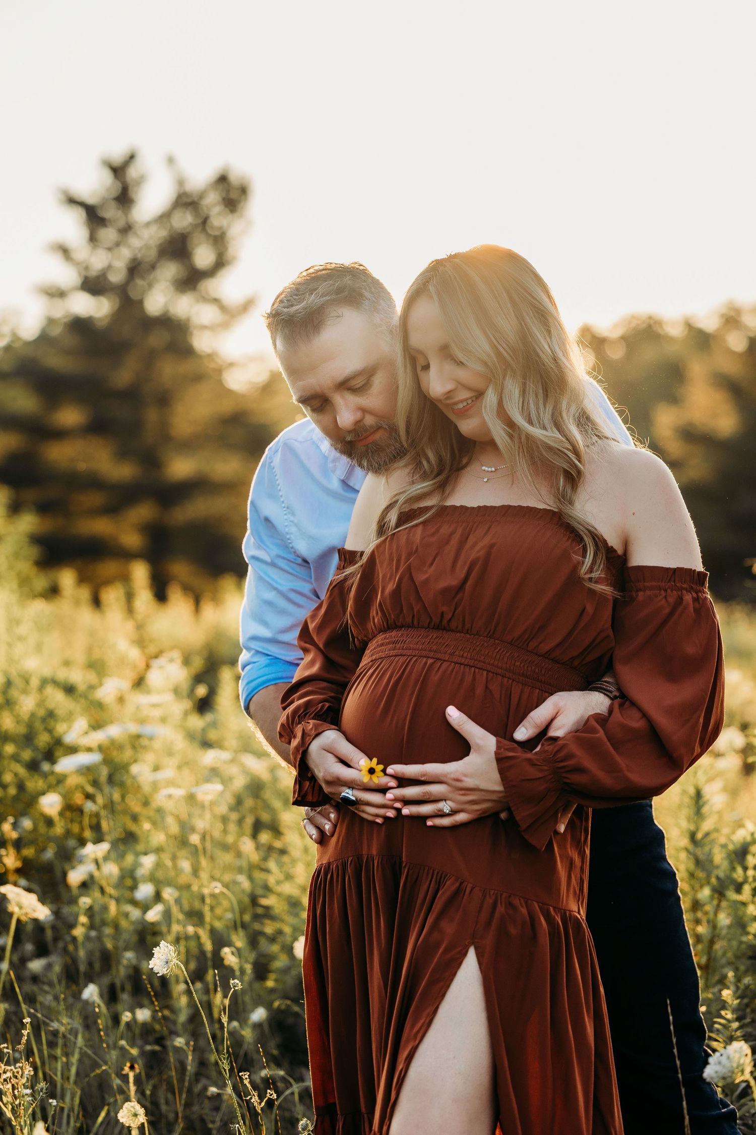 Couple in golden field, pregnant woman in rust off-shoulder gown, man embracing her from behind at sunset.