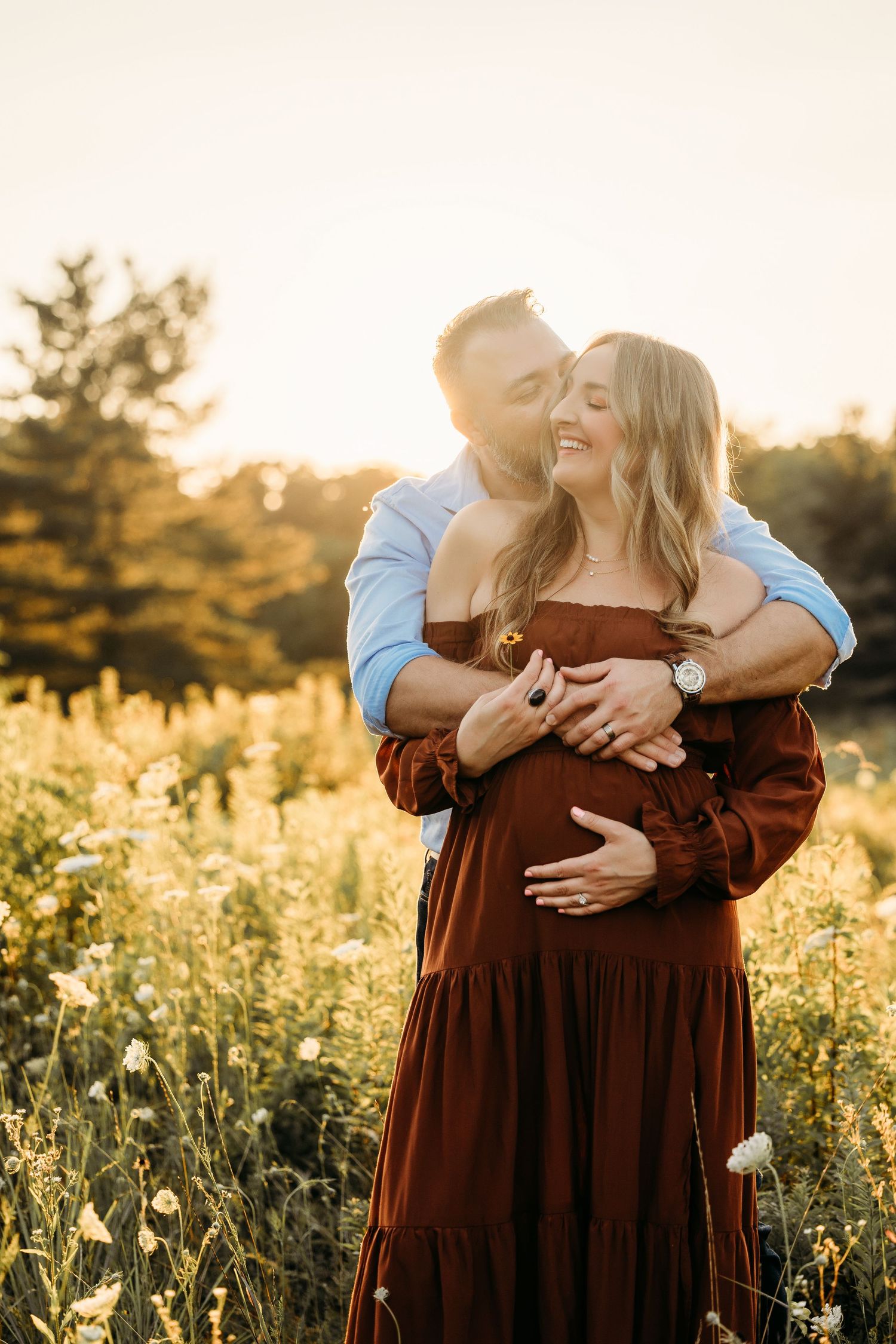 Couple embracing in golden sunflower field at sunset, man kissing pregnant woman in burgundy dress.