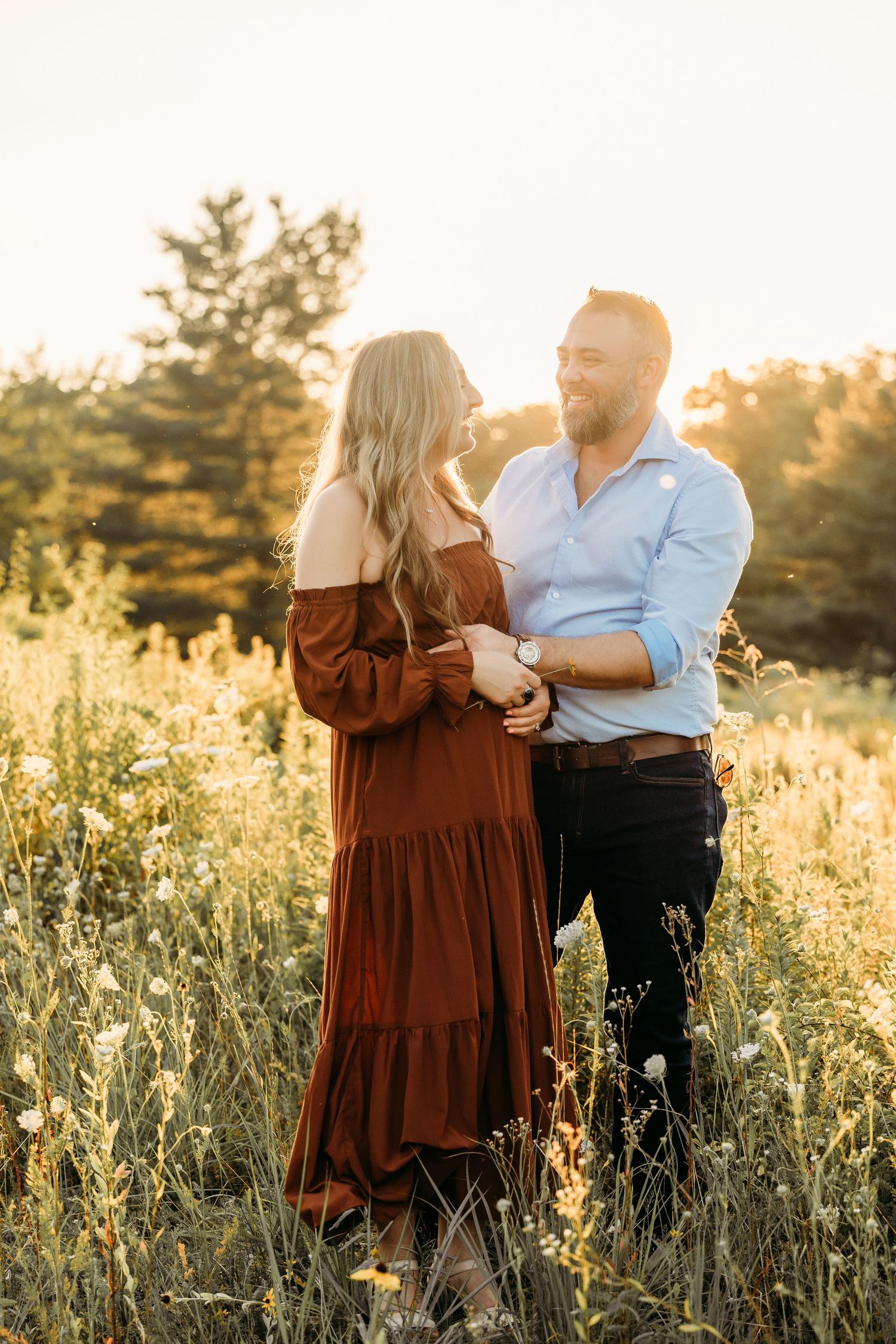 A couple embraces in a sunlit wildflower field, the woman in a rust dress and man in blue shirt, golden hour light glowing behind them.