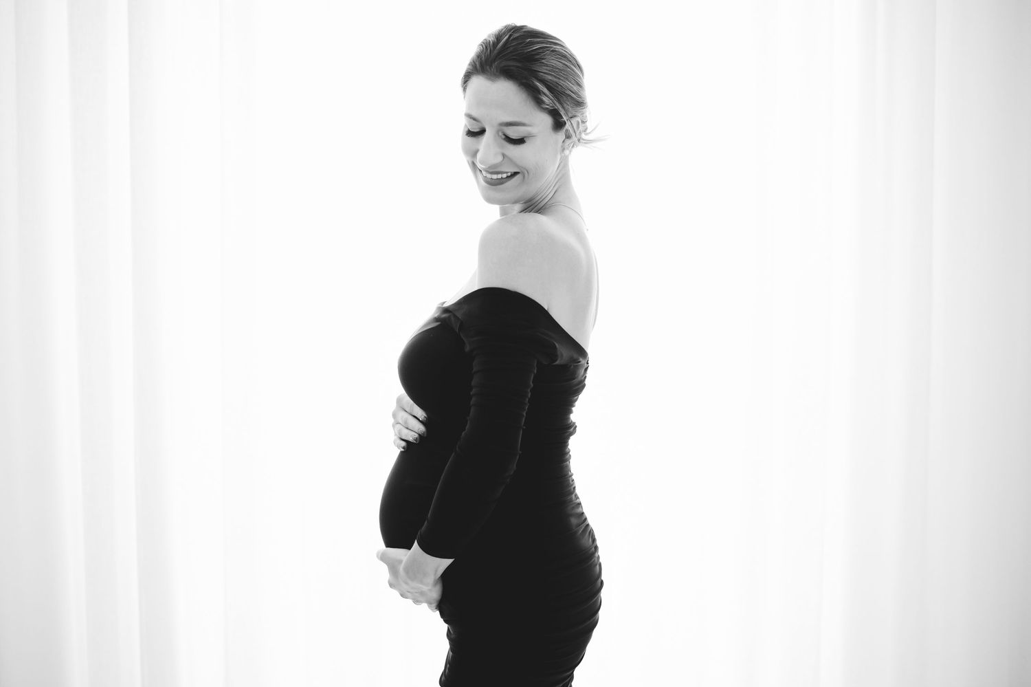Black and white maternity photo of a smiling pregnant woman in a black off-shoulder dress holding her baby bump.