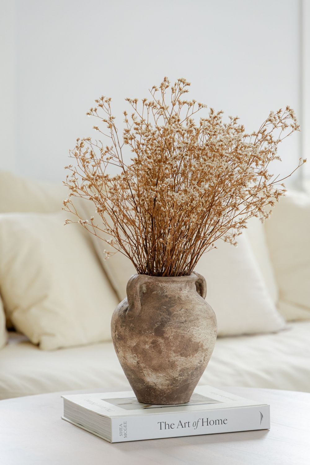 Rustic terracotta vase with dried wildflowers on The Art of Home book, styled on white table near cream sofa.