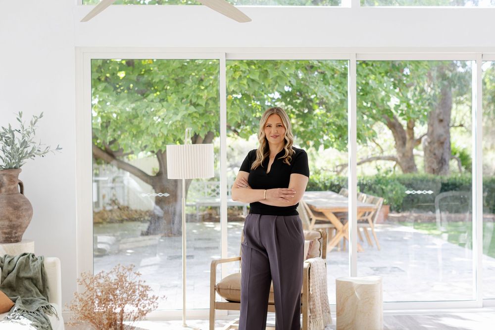 Woman in black top and gray pants stands confidently in a bright, modern sunroom with large windows and lush garden views.