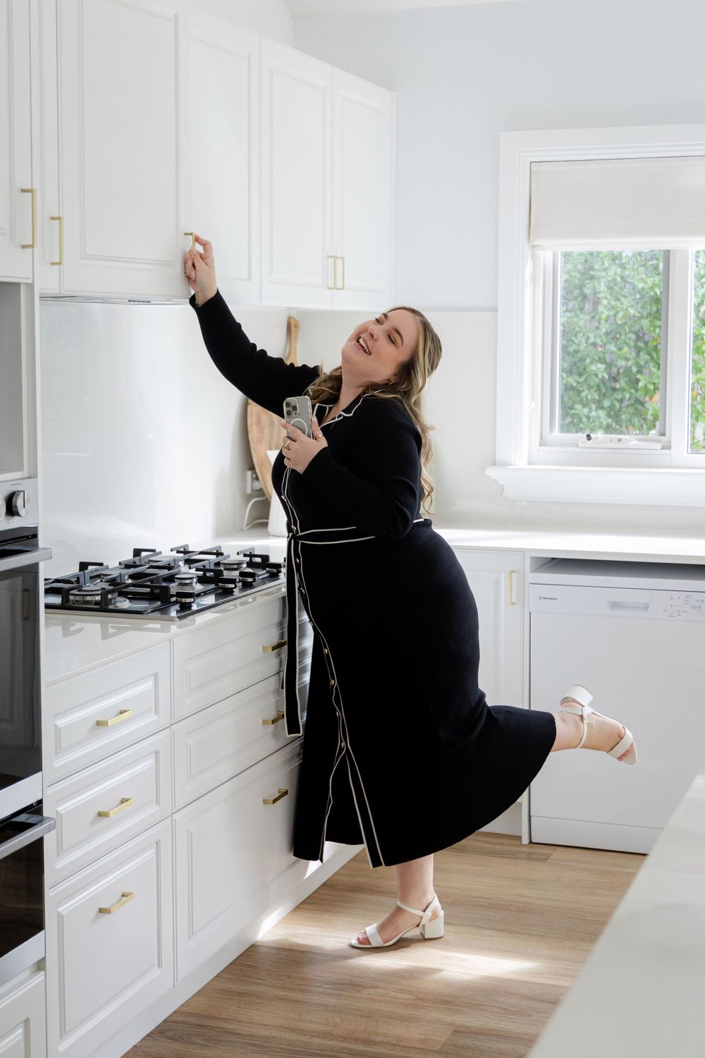 Woman in black maxi dress posing playfully in a modern white kitchen with gold hardware accents.