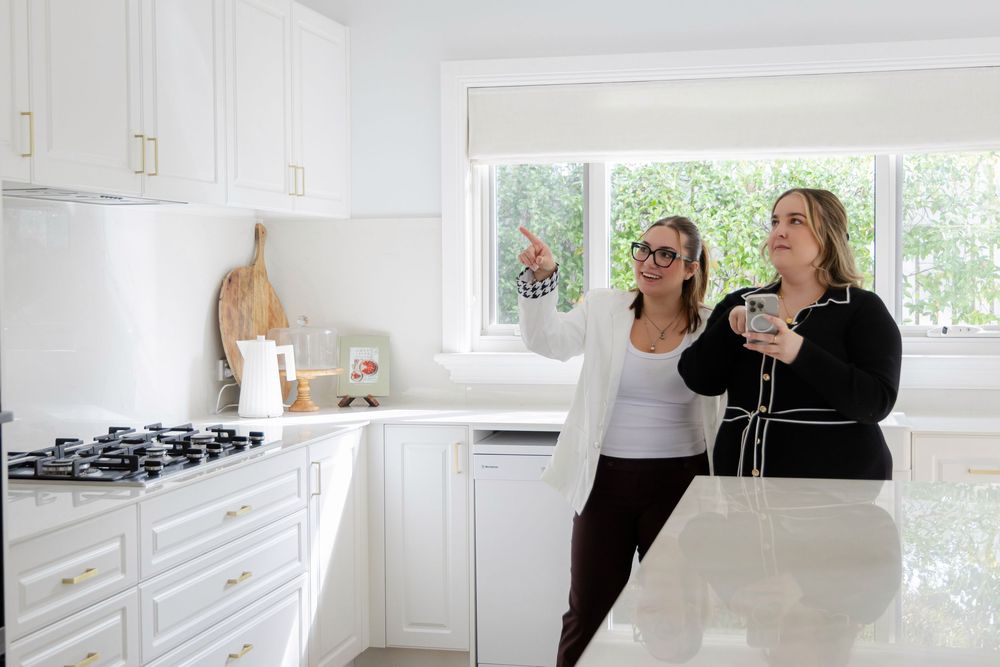 Two women admire a bright, modern white kitchen with marble countertops, white cabinets, and a gas stovetop.