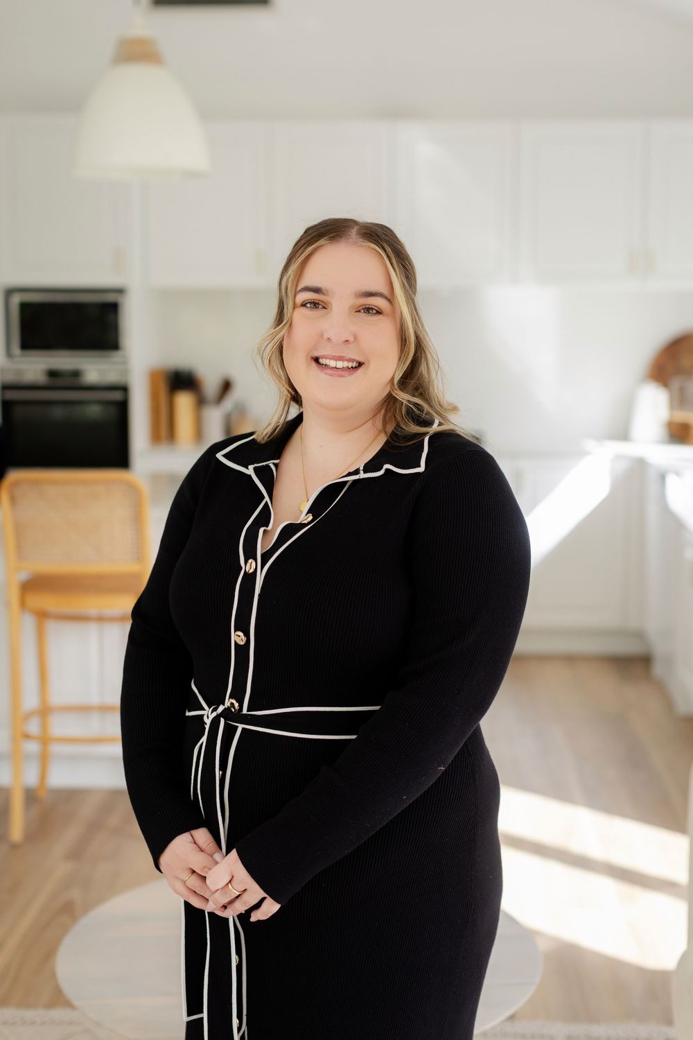 Smiling woman in black dress with white trim stands in bright modern kitchen interior.