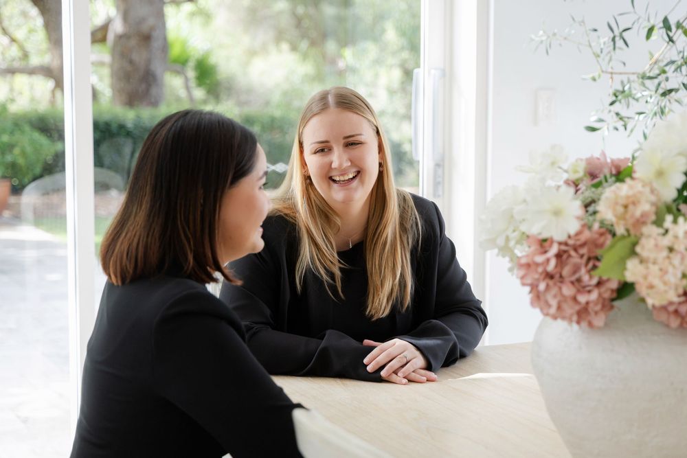 Two women in black tops smile and chat at a marble table with pink floral arrangement nearby.