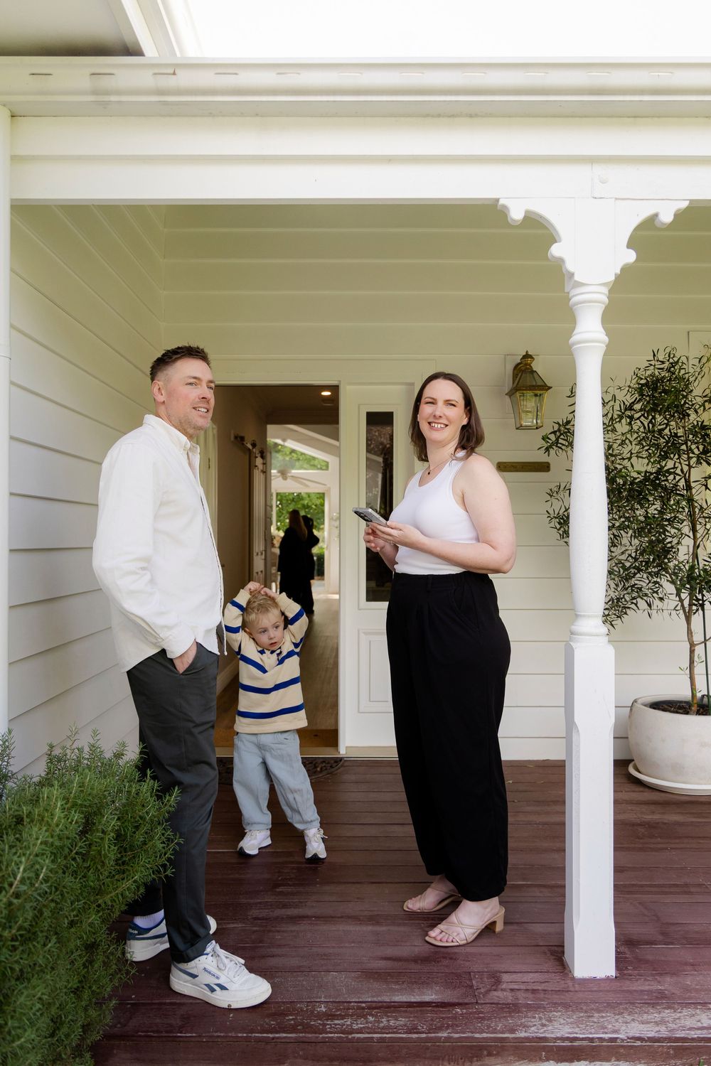 A real estate agent holding a tablet smiles with a man and child on the porch of a charming white weatherboard home.