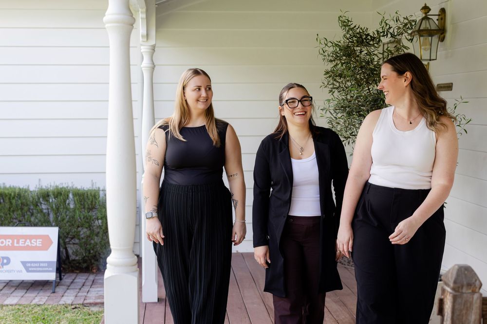 Three women walking together on a porch near a 'For Lease' sign, dressed in elegant black and white outfits.