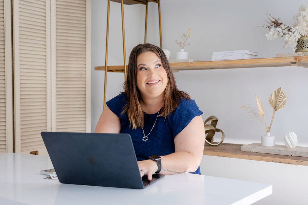 Woman in blue top smiling at desk with laptop in bright modern home office with wooden shelving.