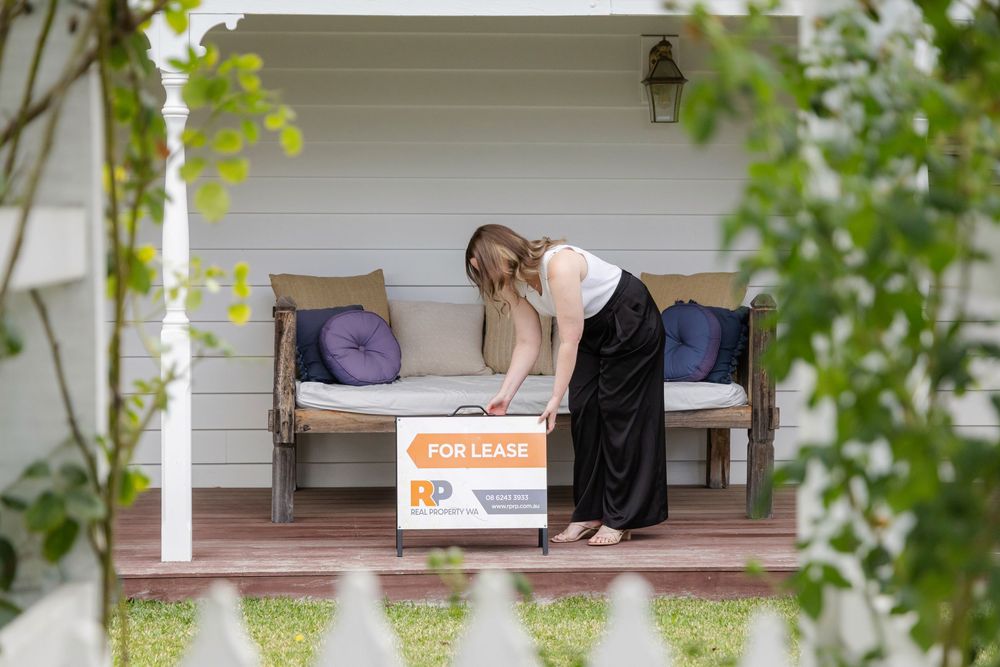 Woman placing a 'For Lease' real estate sign on a porch of a charming white weatherboard home.