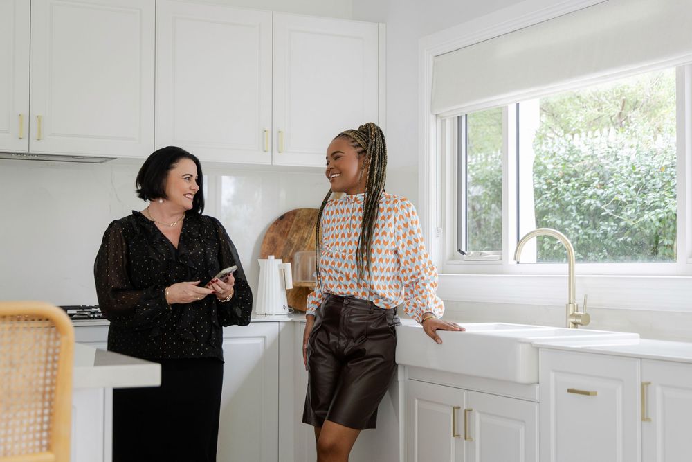 Two women chatting in a modern white kitchen, one holding a tablet, standing near bright windows.
