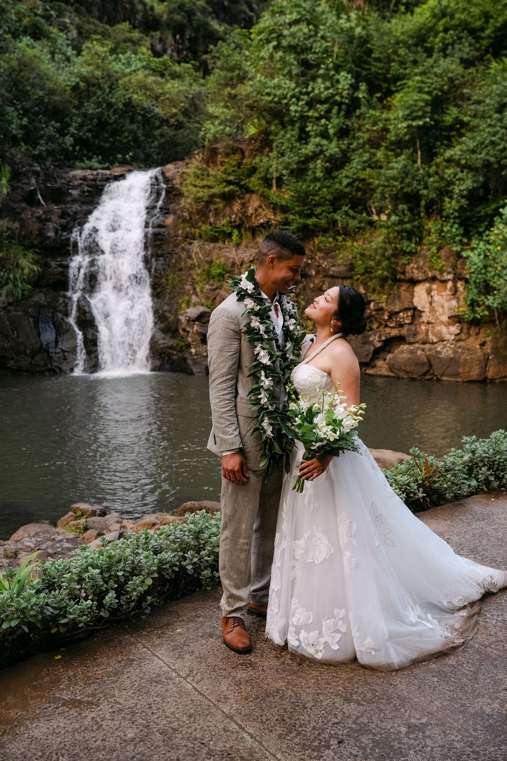 Bride and groom share a romantic moment in front of a lush waterfall in Hawaii, wearing floral leis.