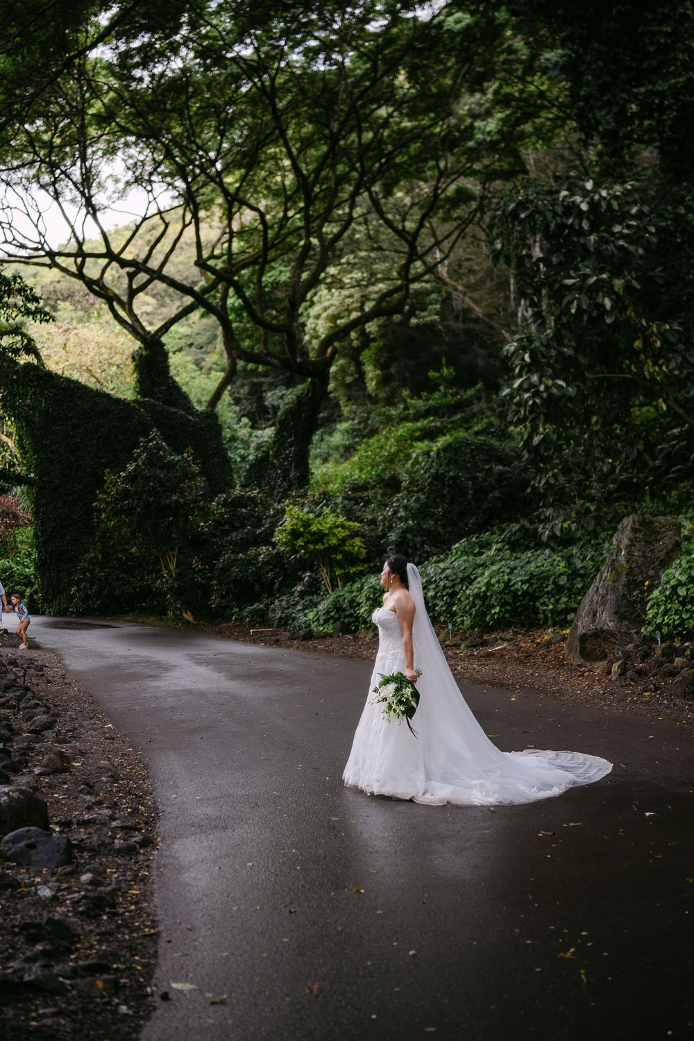 Bride in white gown and veil holding bouquet stands on a winding path surrounded by lush green trees.