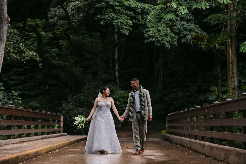 Bride and groom hold hands on a wooden bridge surrounded by lush green forest on their wedding day.