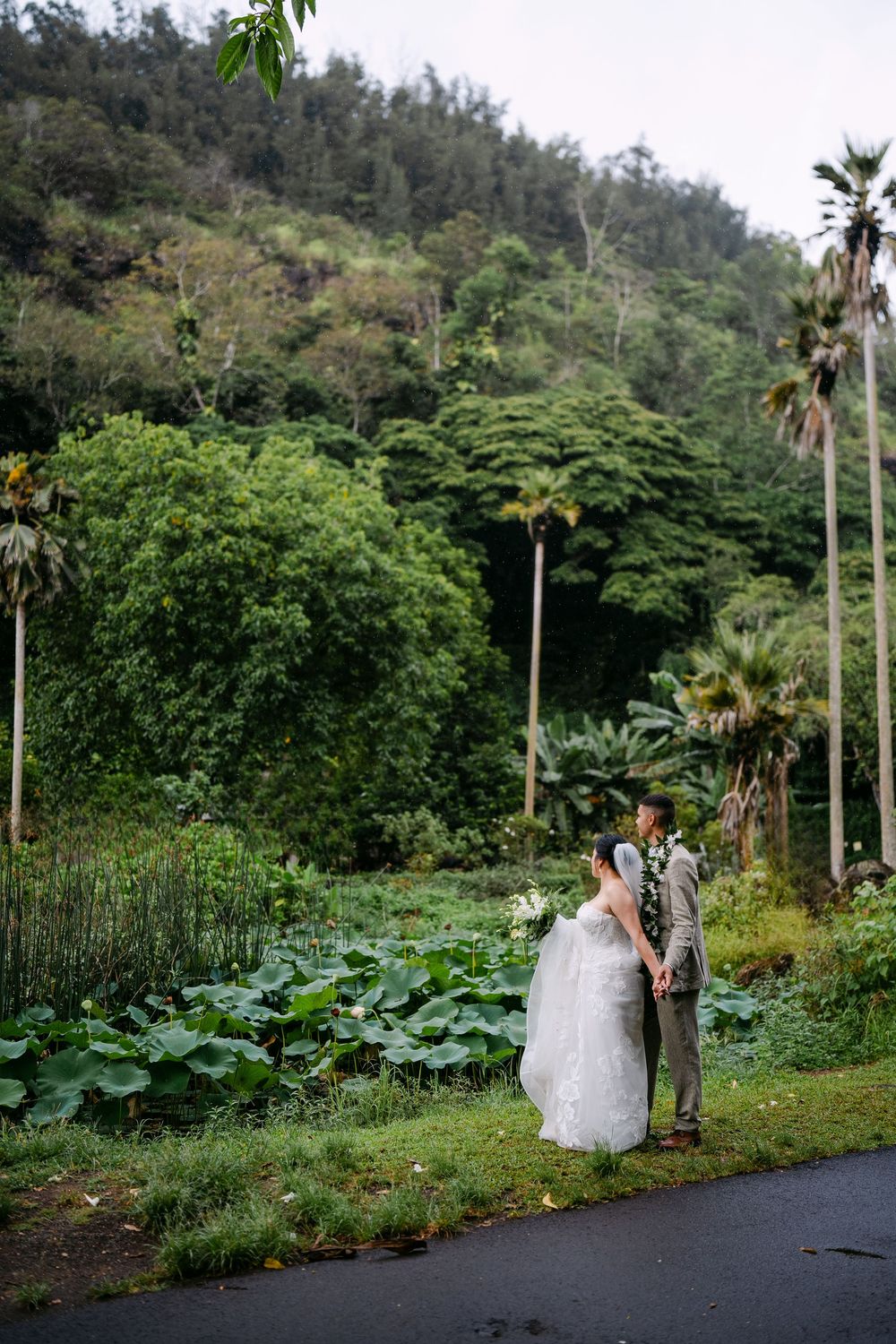 Bride and groom hold hands in lush tropical garden with palm trees and green hillside backdrop on their wedding day.