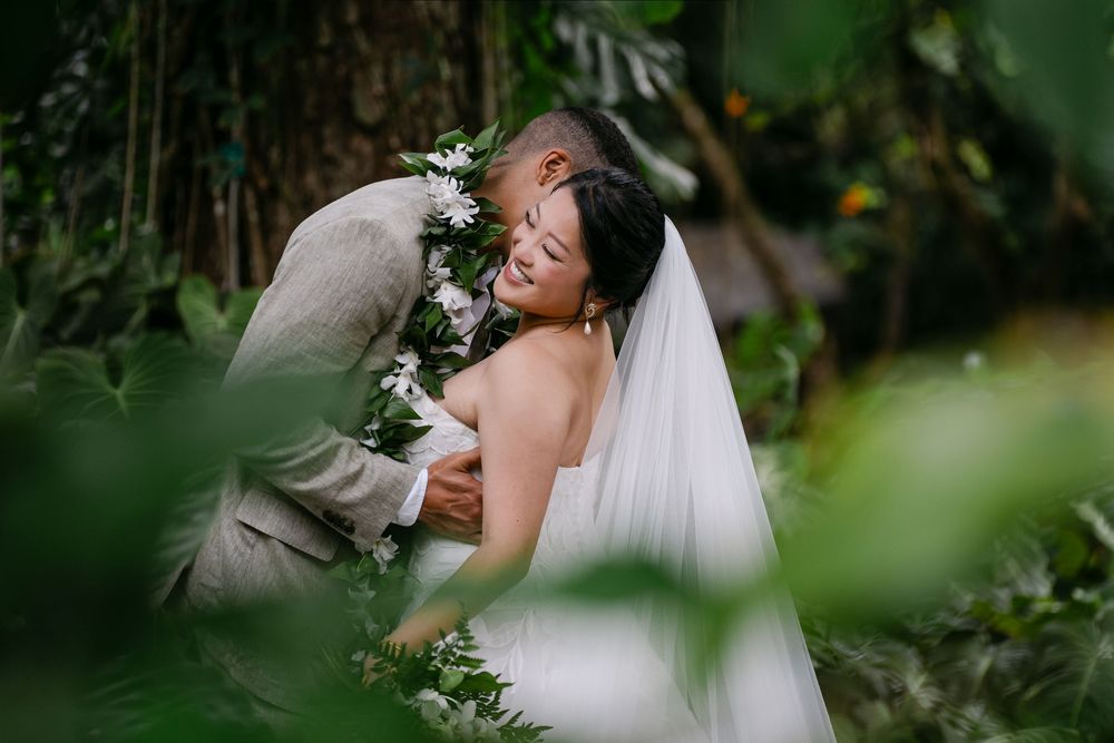 Bride and groom embrace tenderly in a lush tropical garden, bride wearing a white gown and veil with floral lei.