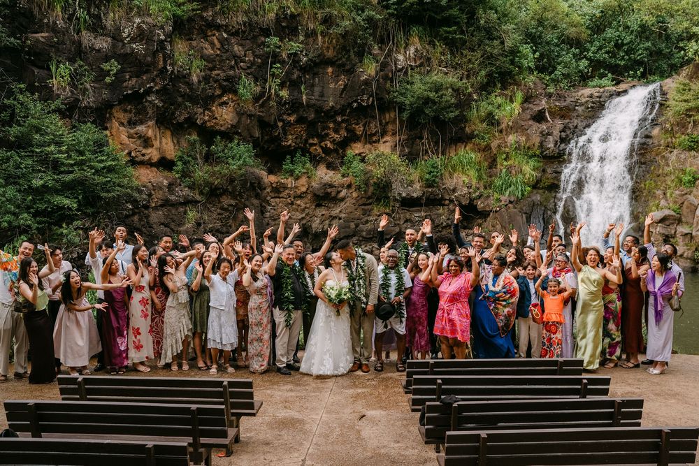 Wedding group photo with guests cheering in front of a stunning waterfall and lush greenery outdoor ceremony.