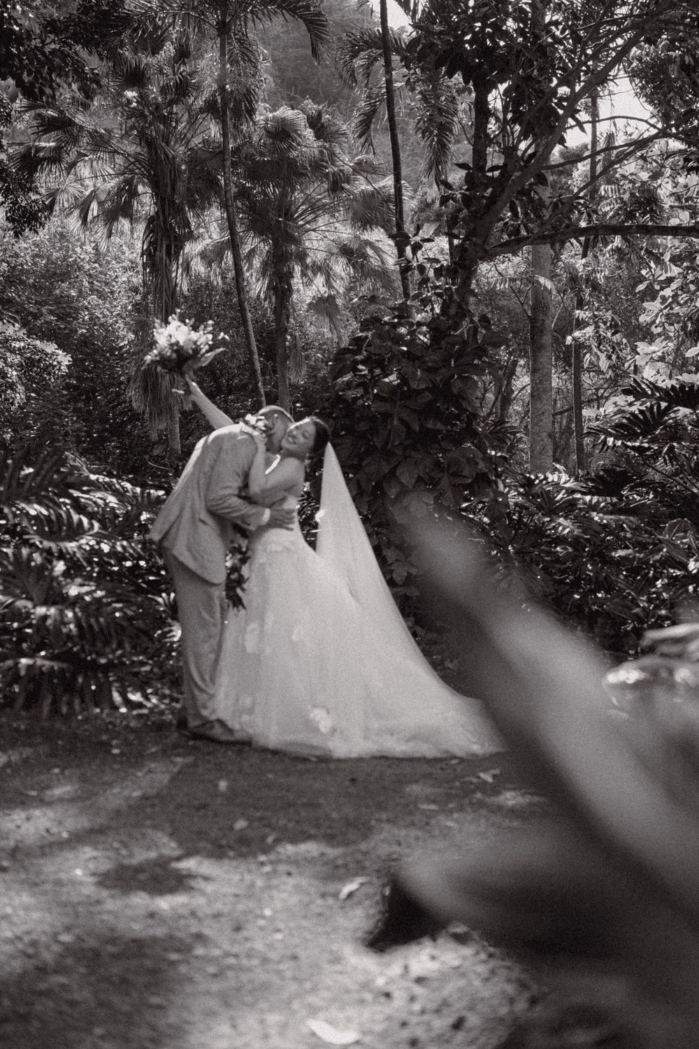Black and white photo of a couple sharing a romantic dip kiss in a lush garden, bride in flowing gown holding bouquet.