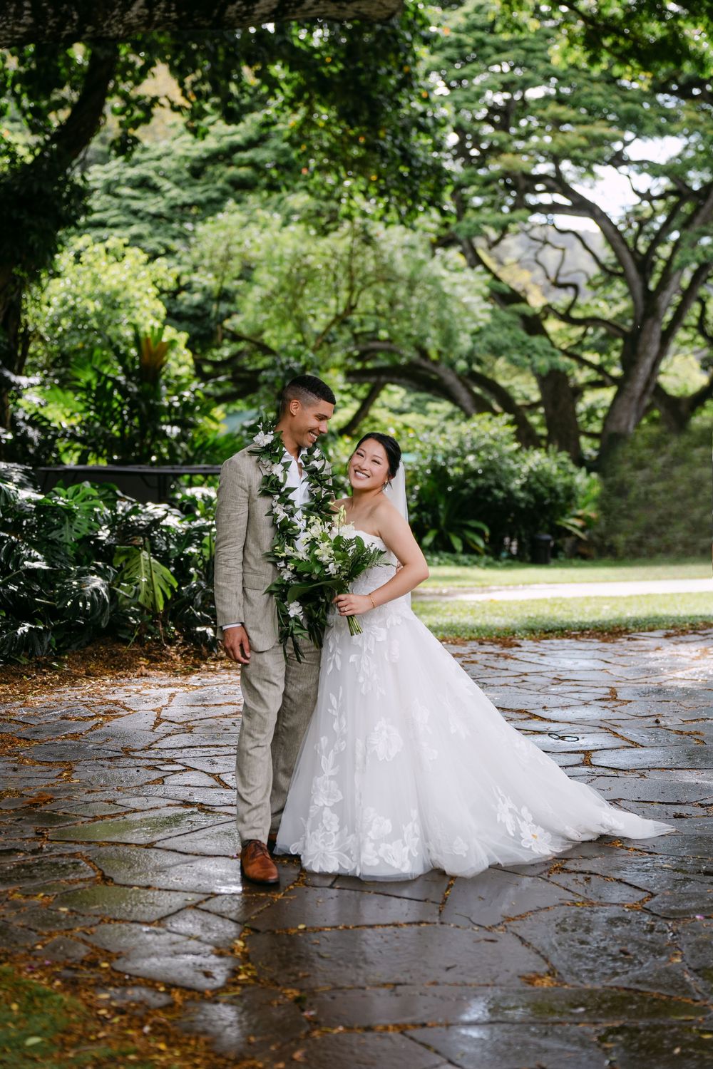 Bride and groom embrace on wet stone path surrounded by lush green trees, bride holding white floral bouquet.