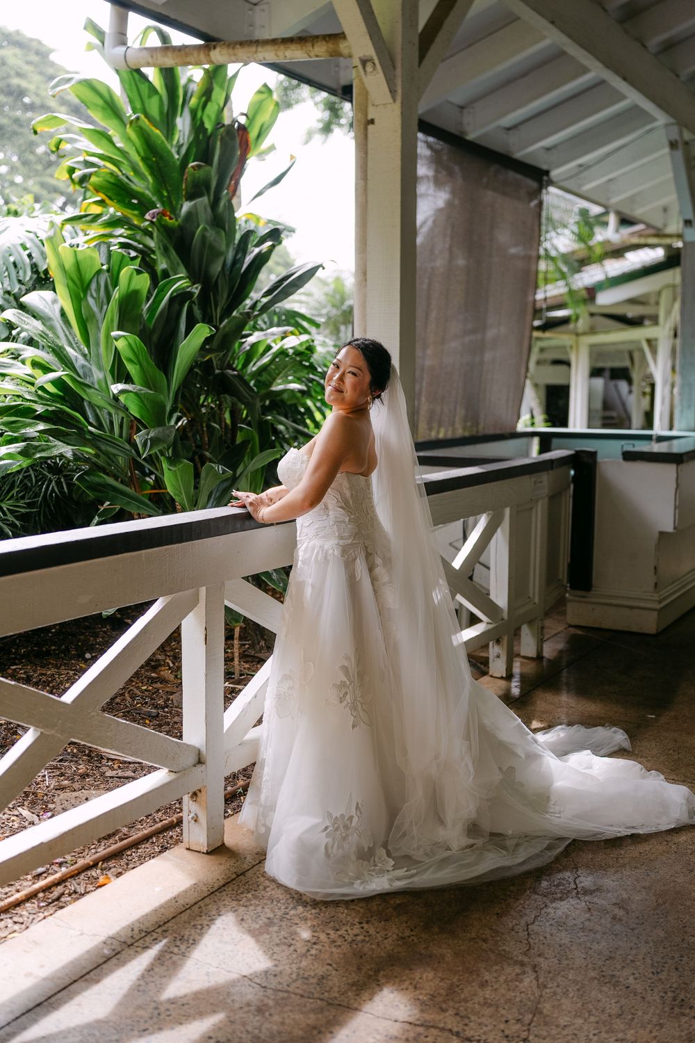Bride in strapless lace gown with veil stands on tropical venue balcony surrounded by lush greenery.