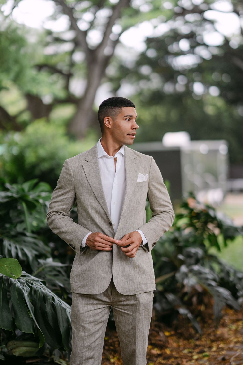 A well-dressed man in a beige linen suit adjusts his jacket buttons while standing outdoors among lush green tropical plants.