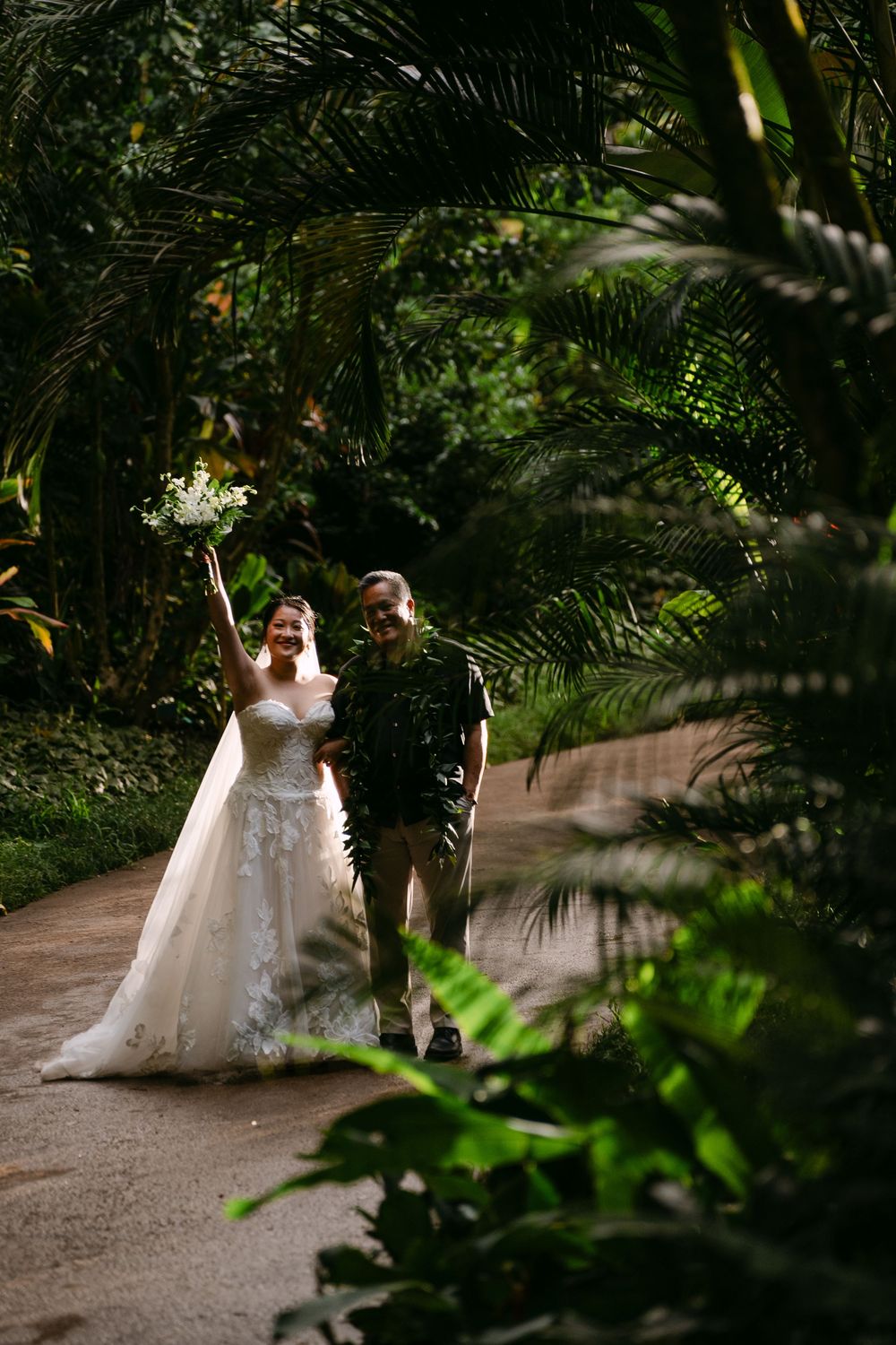 Bride in white gown raises bouquet joyfully beside groom on lush tropical garden path.