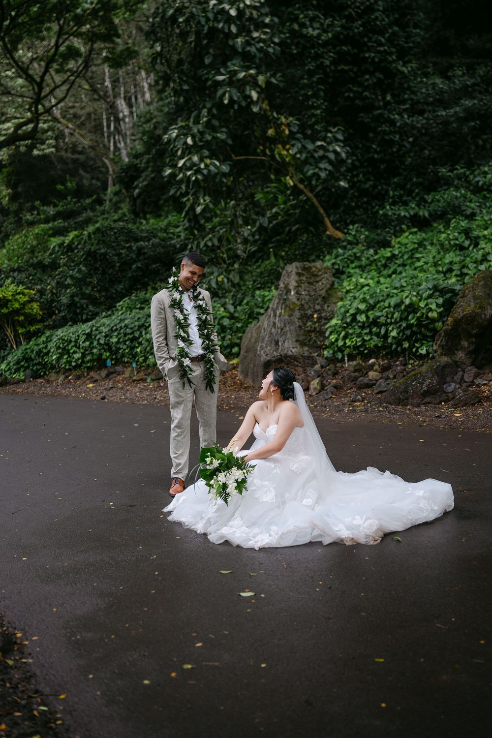 Bride in white gown sits on wet pavement holding bouquet while groom in tan suit with lei stands nearby in lush garden.