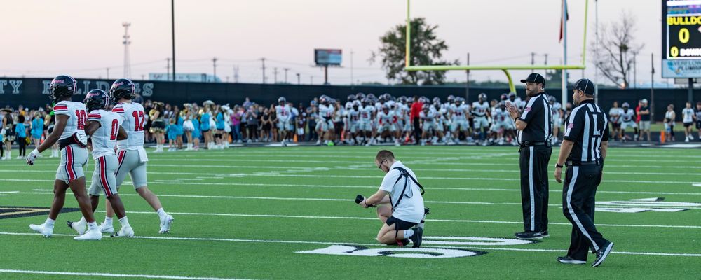 Sports photographer kneels on football field sideline while referee and players stand nearby.