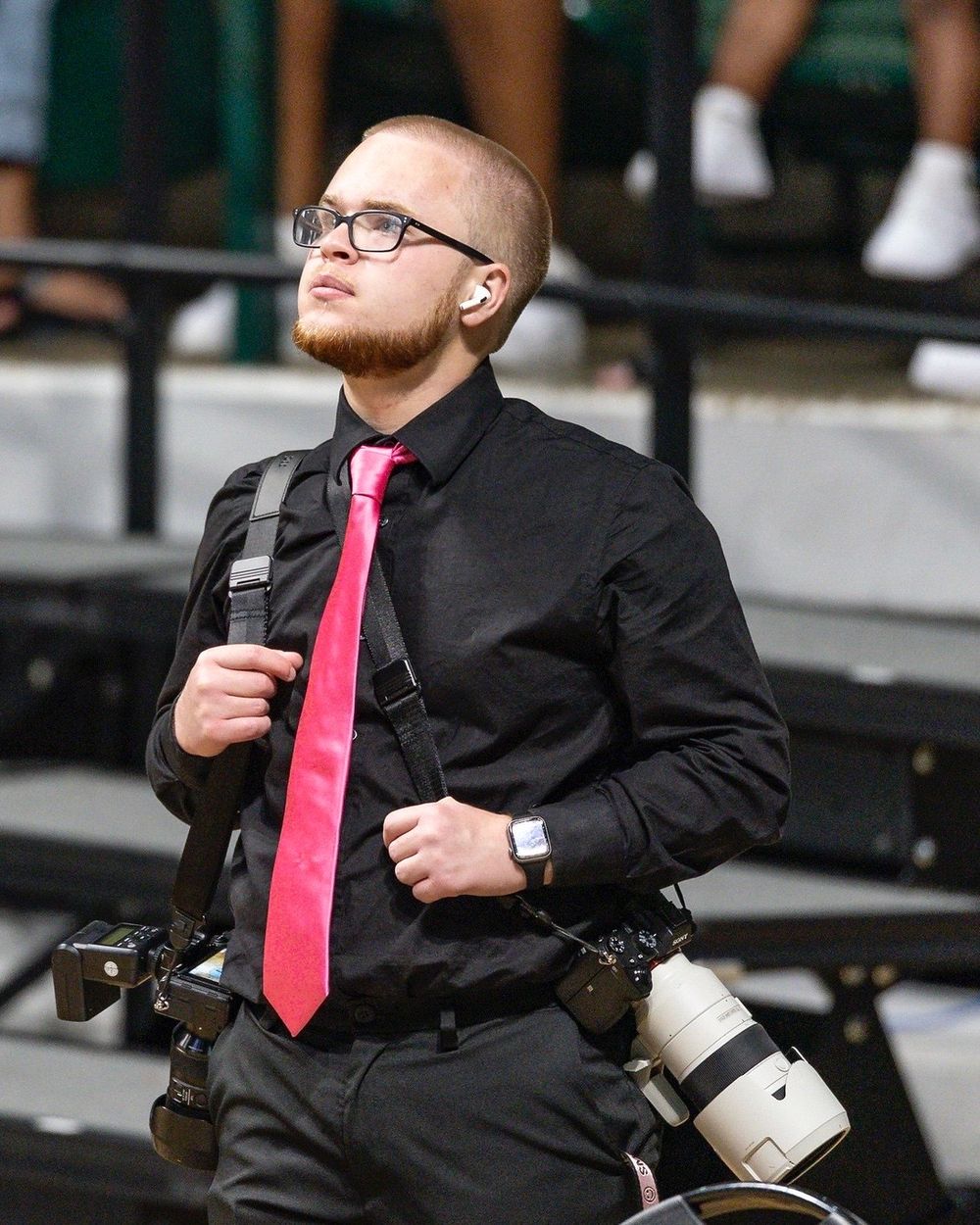 Professional sports photographer in black shirt and pink tie carries dual cameras at sporting event.